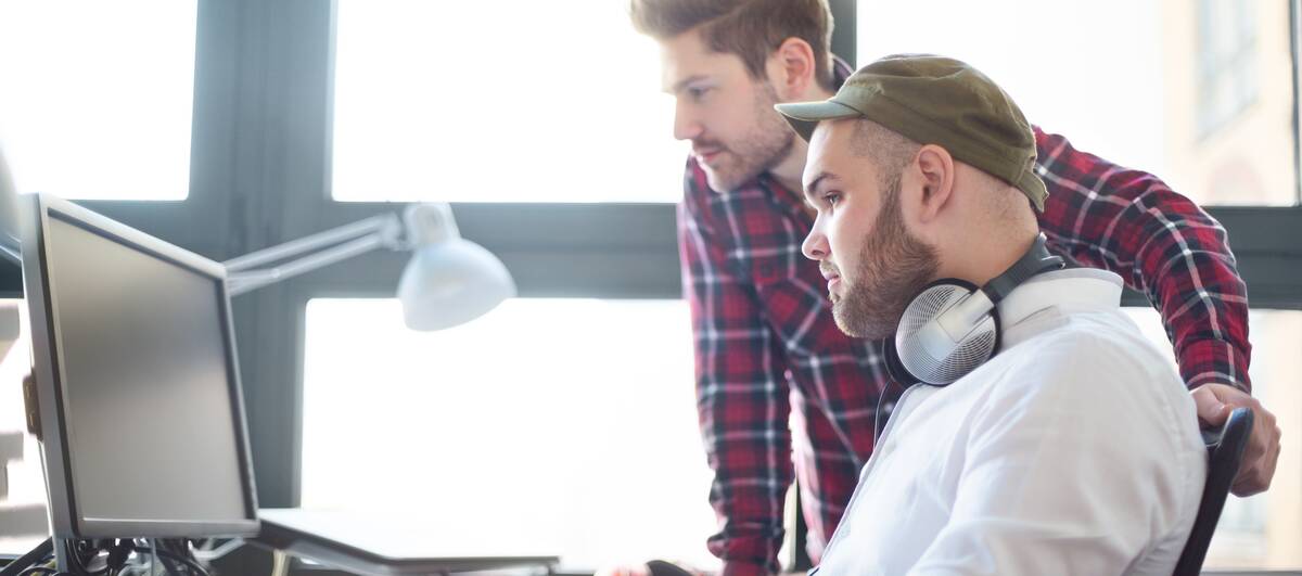 Zwei Männer arbeiten gemeinsam an einem Computer in einem hellen Büro, einer sitzt, der andere steht dahinter